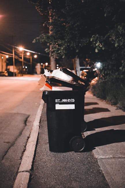 A black wheeled rubbish bin positioned on a residential sidewalk during nighttime, filled with a variety of discarded materials, including paper, cardboard, and plastic items. The bin has a white label on the front with the name 'St. John's' printed on it. The surrounding environment includes illuminated streetlights casting a warm glow over an empty street with trees and hedges along the pavement. The scene suggests potential for private or alternative waste collection services, such as those provided by Waste Clearance Colliers Wood, for disposing of household rubbish. Shadows of the bin extend onto the pavement, and the background features a quiet, darkened street with distant streetlights and minimal traffic, emphasizing an outdoor urban setting suitable for independent rubbish removal arrangements.