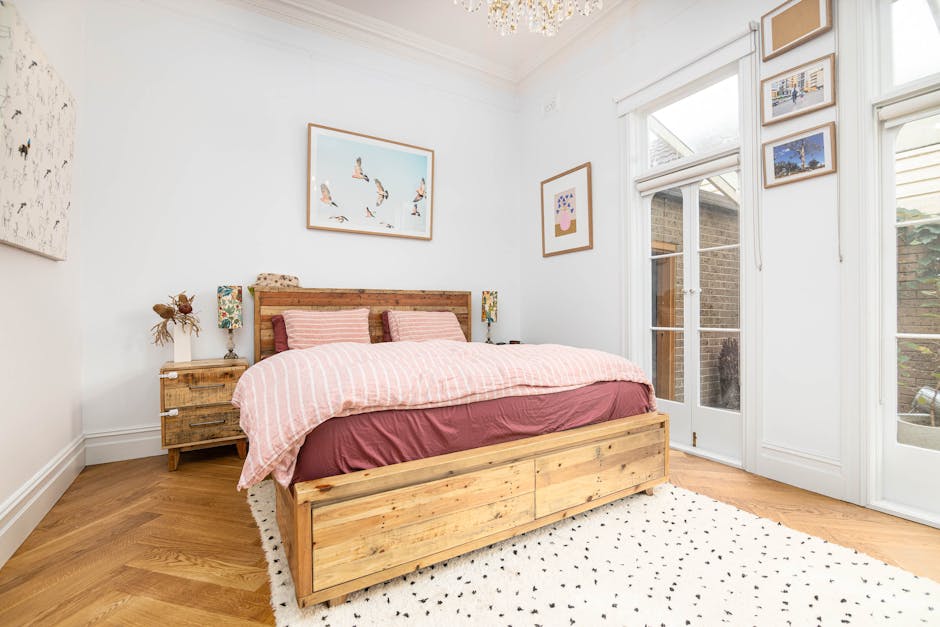 A neatly made double bed with a wooden frame featuring a natural, rustic finish is positioned in the center of a bright, well-lit bedroom. The bed is dressed with a pink and white striped duvet cover and matching pillows, creating a cozy and inviting appearance. Flanking the bed are two small wooden bedside tables, each topped with a patterned table lamp that has a cylindrical lampshade, contributing to warm, ambient lighting. Behind the bed, the wall is decorated with a modern art piece depicting flying birds in soft pastel tones framed in light wood, and other smaller framed images hang on the adjacent wall, adding an aesthetic touch. To the right, a set of white-painted wooden French doors, fitted with glass panels, open onto an outdoor area, revealing brick walls and green foliage beyond. The room’s walls are painted white, complemented by white crown molding and baseboards, and the flooring is warm-toned hardwood. An off-white rug with small black dots is partially visible beneath the bed, adding texture to the space. The overall scene suggests a tidy, stylish bedroom possibly undergoing minimal decluttering, aligning with the principles of professional waste removal and helping maintain a clutter-free environment. Waste Clearance Colliers Wood may assist with private on-site clearance or rubbish collection needs in residential settings like this.