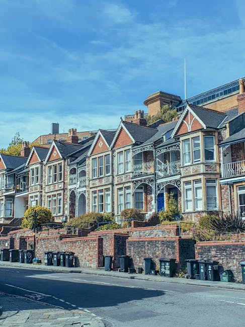 A row of Victorian-style terraced houses with ornate white metalwork on balconies and decorative trim around large bay windows, set on a sloped street with brick retaining walls and low hedges in front. The houses have brick exteriors with some painted sections, and the rooflines feature gabled dormer windows with dark roofing material. In the foreground, there is a paved road with multiple black wheelie bins arranged along the curbside, indicating waste collection points. The setting appears to be a residential area with a clear blue sky overhead, and modern buildings are visible in the background behind the traditional houses. The scene reflects a typical UK urban streetscape where private waste disposal, including refuse collection from residents, is common, supporting independent rubbish removal services such as Waste Clearance Colliers Wood.