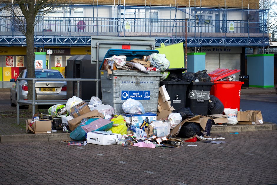 A collection of overflowing waste and rubbish bags accumulated around a large gray mixed waste bin on a paved sidewalk. The waste includes cardboard boxes, paper bags, plastic packaging, and various discarded items in black, white, and colored bags. Some items are spilling onto the surrounding area, which has a metal railing, a parked silver car with a UK license plate, and a background featuring a commercial building with storefronts and a blue scaffolding structure. The scene is outdoors under natural daylight, illustrating a typical urban rubbish disposal area, possibly managed by a service provider such as Waste Clearance Colliers Wood specializing in private waste collection and on-site clearance solutions.