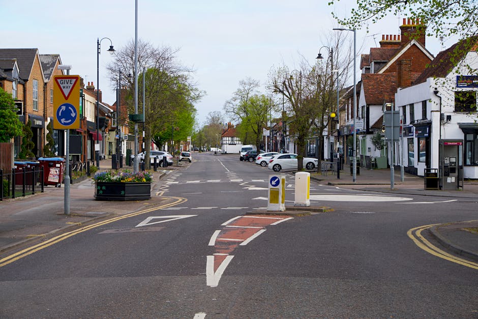 An image of a small urban street scene showing a paved intersection with a central traffic island that has white and yellow bollards and a blue circular sign indicating a one-way direction. Surrounding the street are commercial buildings, consisting of brick and white-painted facades with large windows and signage, some with awnings. On the left side, there are residential-style houses with small front gardens and flower boxes, as well as a recycling bin. On the right side, a parking meter and street lamps line the sidewalk, which features trees with light green foliage and some leafless branches. The sky appears overcast with diffused lighting, and several parked cars are visible along the street, suggesting regular urban activity. The scene captures a quiet moment in a typical UK high street environment, illustrating an area potentially suitable for private waste collection or rubbish clearance services like those provided by Waste Clearance Colliers Wood, especially in the context of maintaining cleanliness in retail and residential zones.