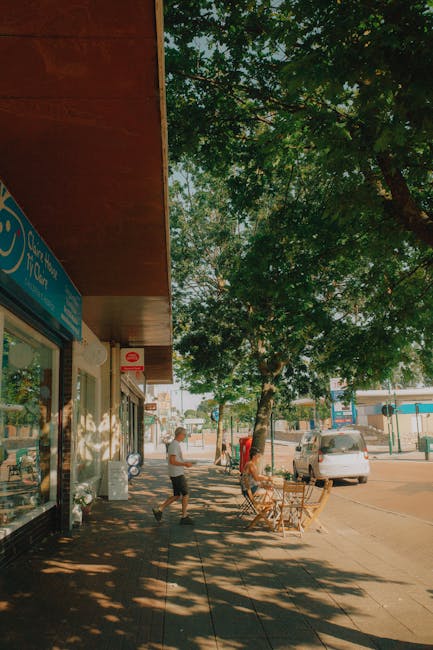 A wide view of a quiet urban sidewalk in Colliers Wood, featuring a shopfront on the left with a blue sign and large display window, partially shaded by a wooden awning. In the foreground, a person is seen walking past outdoor seating that includes two wicker chairs and a small table, positioned beneath a large leafy tree providing dappled shade. Further down the pavement, another individual stands near a trash bin, with additional pedestrians visible in the distance. Parked cars line the curb on the right side of the street, and the background shows a mix of low-rise buildings and trees, suggesting a typical commercial and residential area. The scene is illuminated by natural daylight, with sunlight filtering through the tree’s green foliage, casting shadows on the pavement, creating a calm and everyday atmosphere consistent with local rubbish removal or waste management services occasionally supporting private disposal solutions in the area. Waste Clearance Colliers Wood is aware of the need for efficient rubbish clearance within busy high street environments, although no equipment or waste is visible in this particular image.