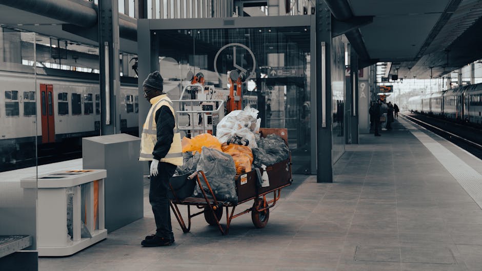 A man dressed in a dark jacket, black pants, and wearing a knitted cap and high-visibility vest stands on a modern train station platform next to a large cart filled with various waste bags and discarded items, including black, grey, and orange rubbish bags, some appearing partially open or overfilled, with loose debris visible. The cart has a metal frame and large wheels, positioned close to the station’s glass-walled lift entrance and modern metal awnings, while a train with red and white doors is parked on the left side of the platform. In the background, there are a few other passengers and the train tracks extending into the distance under an overcast sky. The scene appears to capture an instance of private waste removal or rubbish clearance, possibly arranged through services like Waste Clearance Colliers Wood, at a contemporary, urban train station environment suited for on-site or independent rubbish disposal activities.